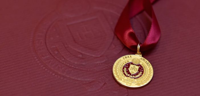 A close-up of the gold medal given to awardees, against a maroon background.