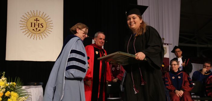 A girl wearing a black graduation gown smiles and receives a plaque from a woman wearing a light blue graduation gown