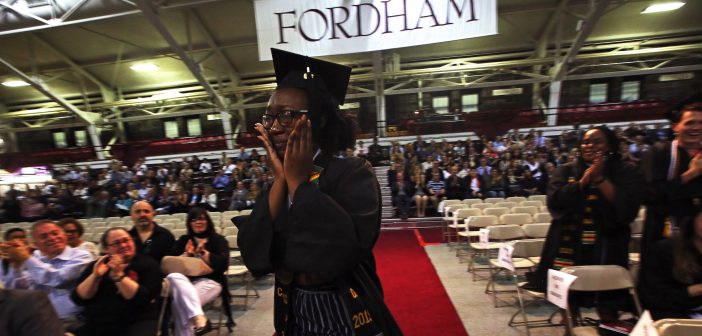 A girl wearing a black graduation gown holds hands to her tear-streaked face and walks along an aisle