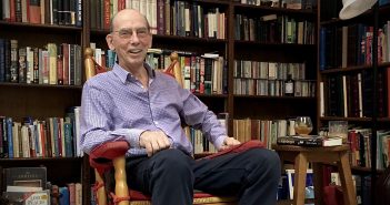 A smiling elderly man sitting in front of a row of bookshelves
