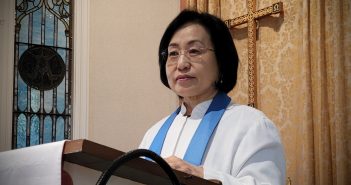 An Asian-American woman wearing glasses and a white pastor's robe stands in front of a pulpit.