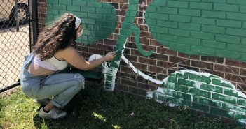 A girl wearing overalls and a mask squats in front of a brick mural and paints with a green brush.