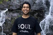 A man wearing a black t-shirt that says "Soul of Fordham" smiles in front of a waterfall.