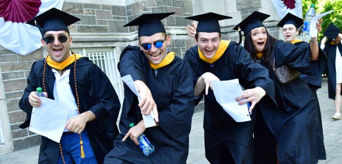 Four students wearing black graduation gowns make silly poses and point at the camera.