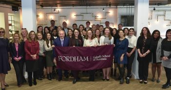 A large group of people stand and smile while holding a maroon flag that says "FORDHAM Gabelli School Business."