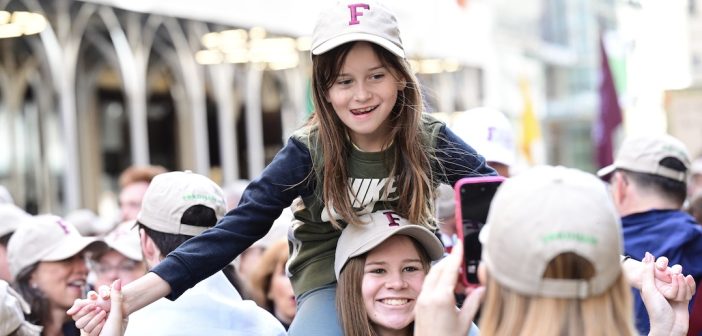 A girl wearing a Fordham baseball cap sits on the shoulders of a young woman wearing a Fordham baseball cap.